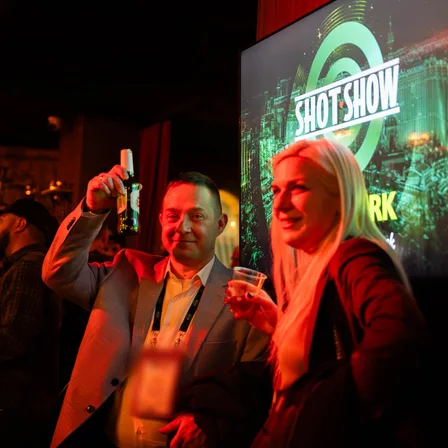 Two guests with drinks at an evening trade-show reception, a SHOT Show screen behind them