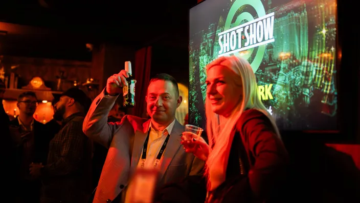 Two guests with drinks at a cocktail reception, one raising a bottle to the camera, an event screen lit behind them.