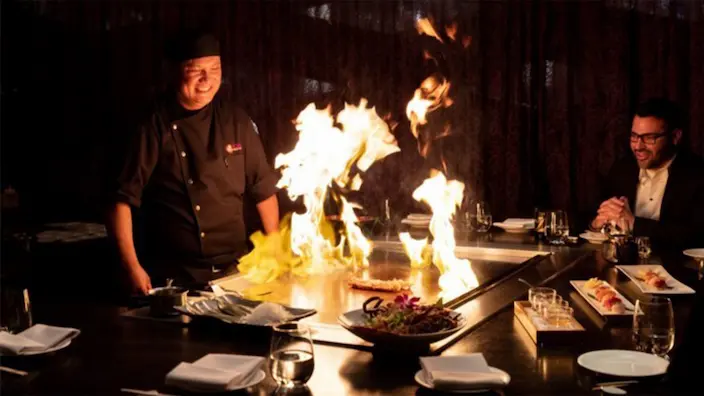 A chef cooking at a teppanyaki table with guests seated around it, flames rising from the grill.